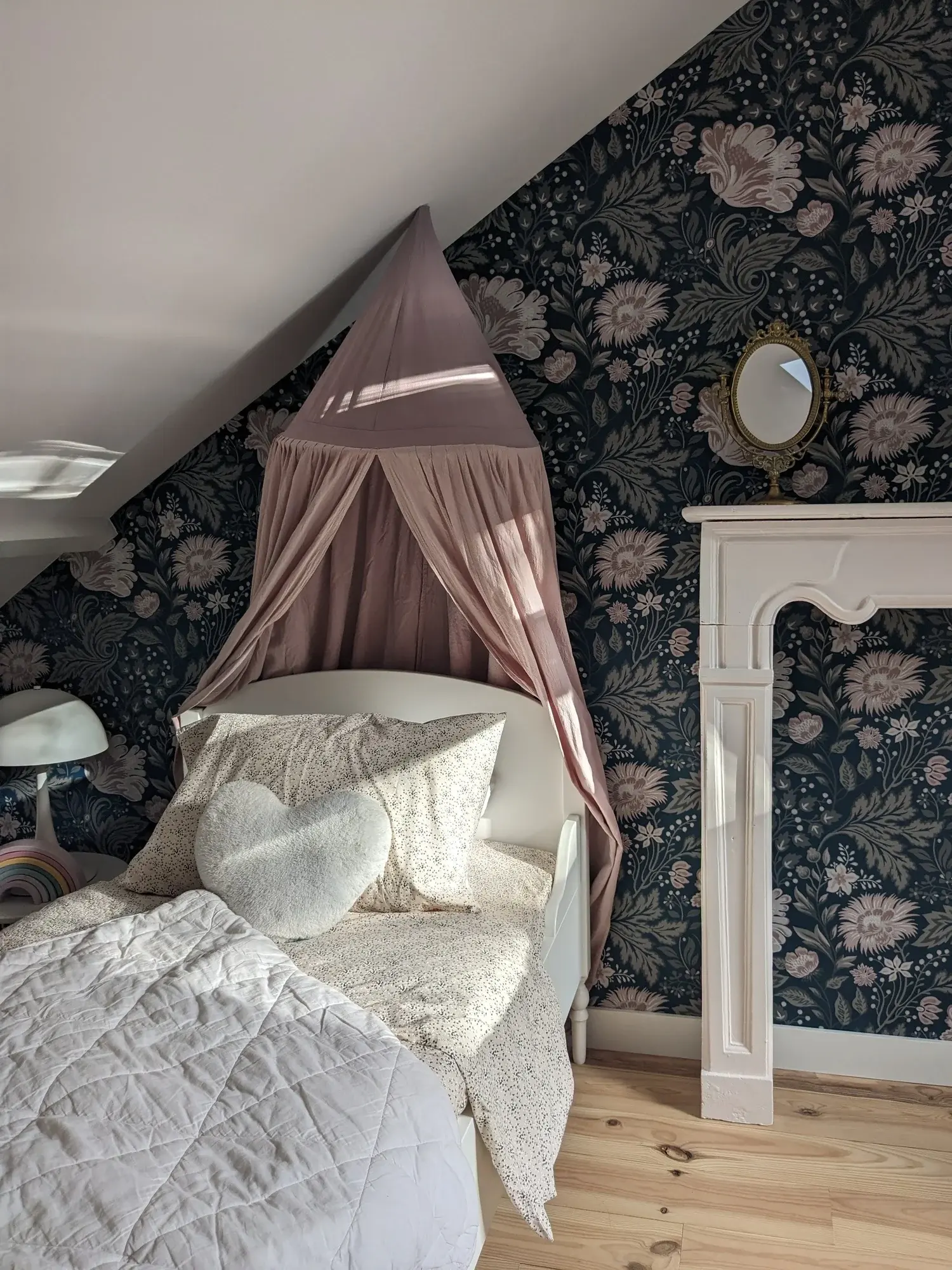 Chambre d’enfant mansardée, papier peint fleuri bleu nuit et ciel de lit rose poudré